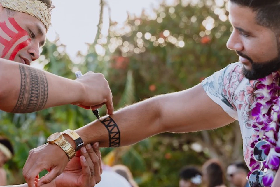 A person with face paint applies a tribal-style temporary tattoo to another person's arm outdoors in a tropical setting.