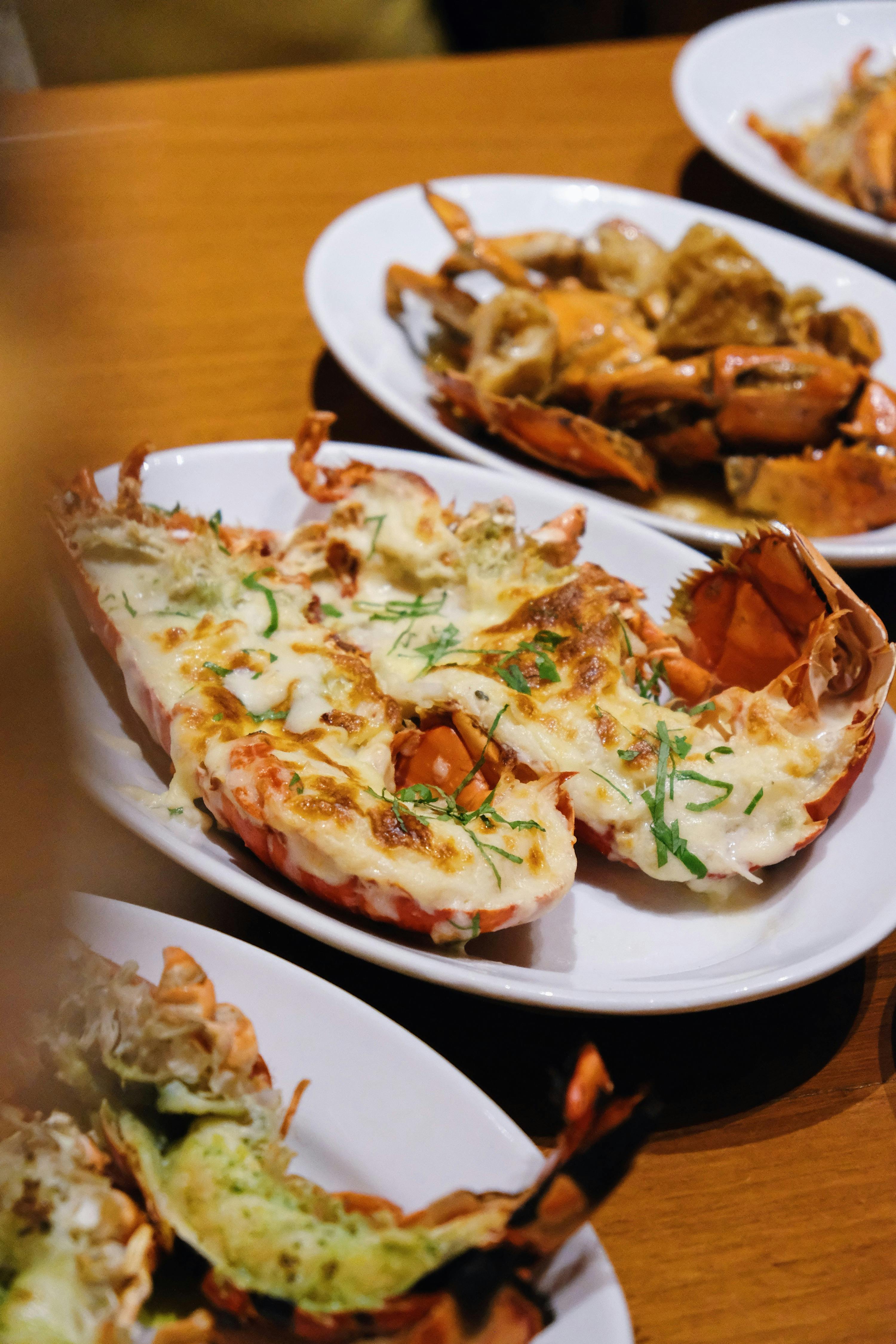 Plates of grilled seafood, including lobsters topped with melted cheese and herbs, served on a wooden table.