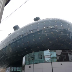 Modern byggnad med en reflekterande, mörkblå böjd fasad med många metallfästen, i anslutning till en traditionell struktur.