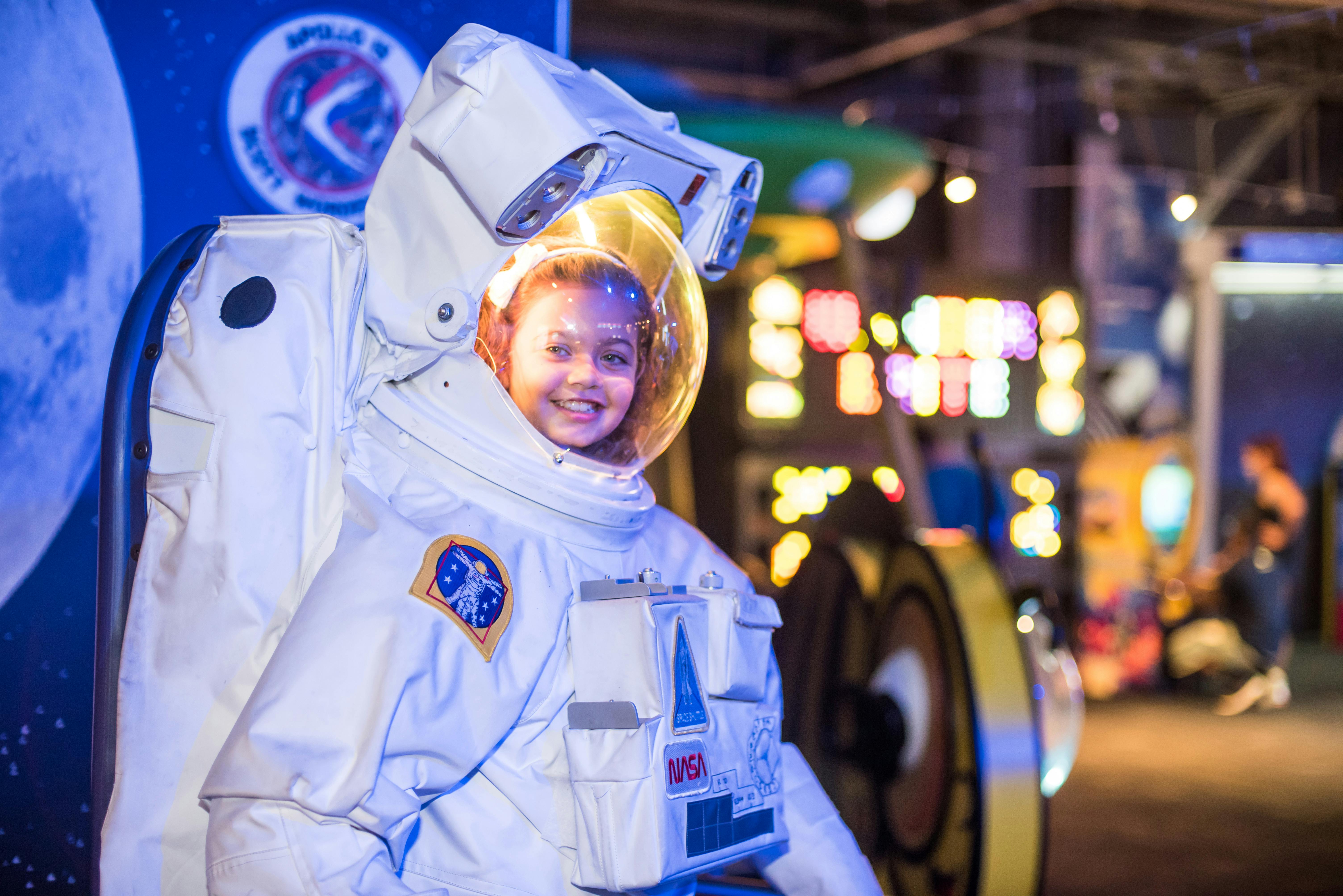 A child smiling inside a large white astronaut suit replica, with colorful lights and displays in the background.