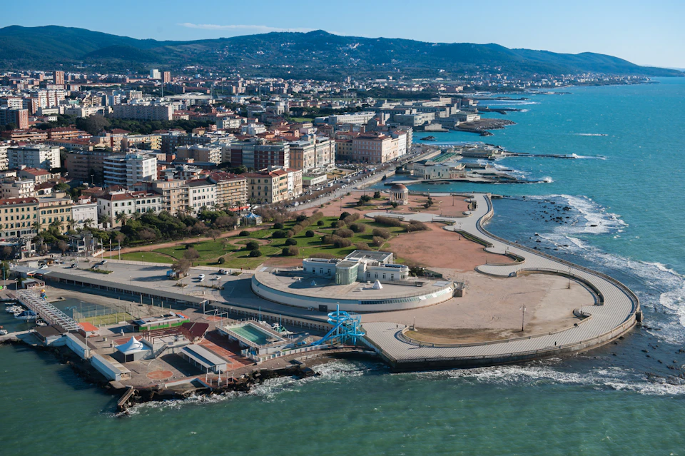 Aerial view of a coastal city with buildings, roads, parks, and a shoreline along a turquoise sea bordered by a promenade.