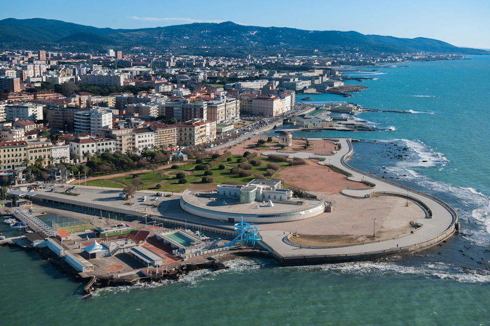 Aerial view of a coastal city with buildings, roads, parks, and a shoreline along a turquoise sea bordered by a promenade.