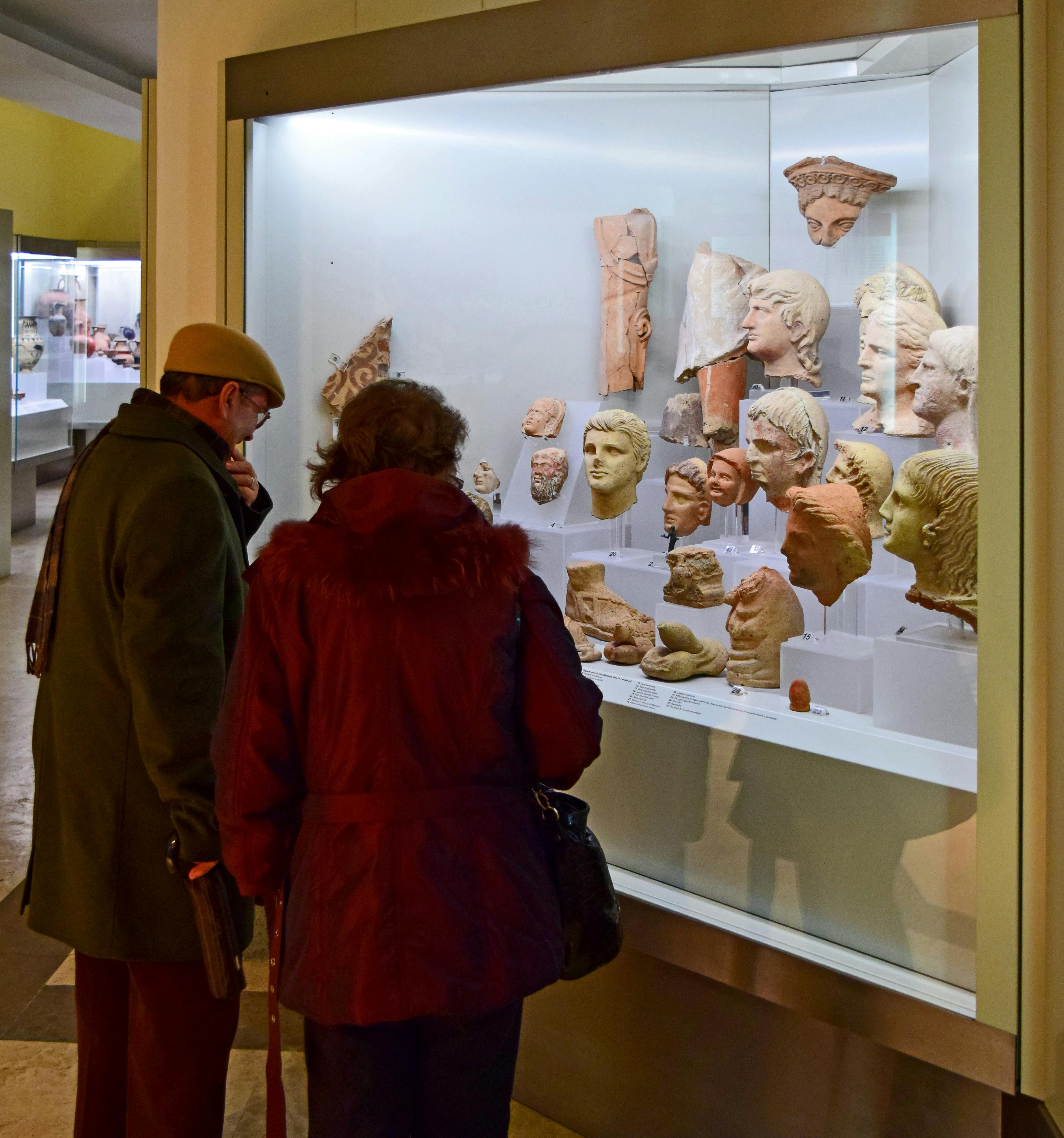 Twee mensen bekijken een museumvitrine met verschillende oude gebeeldhouwde hoofden en fragmenten.