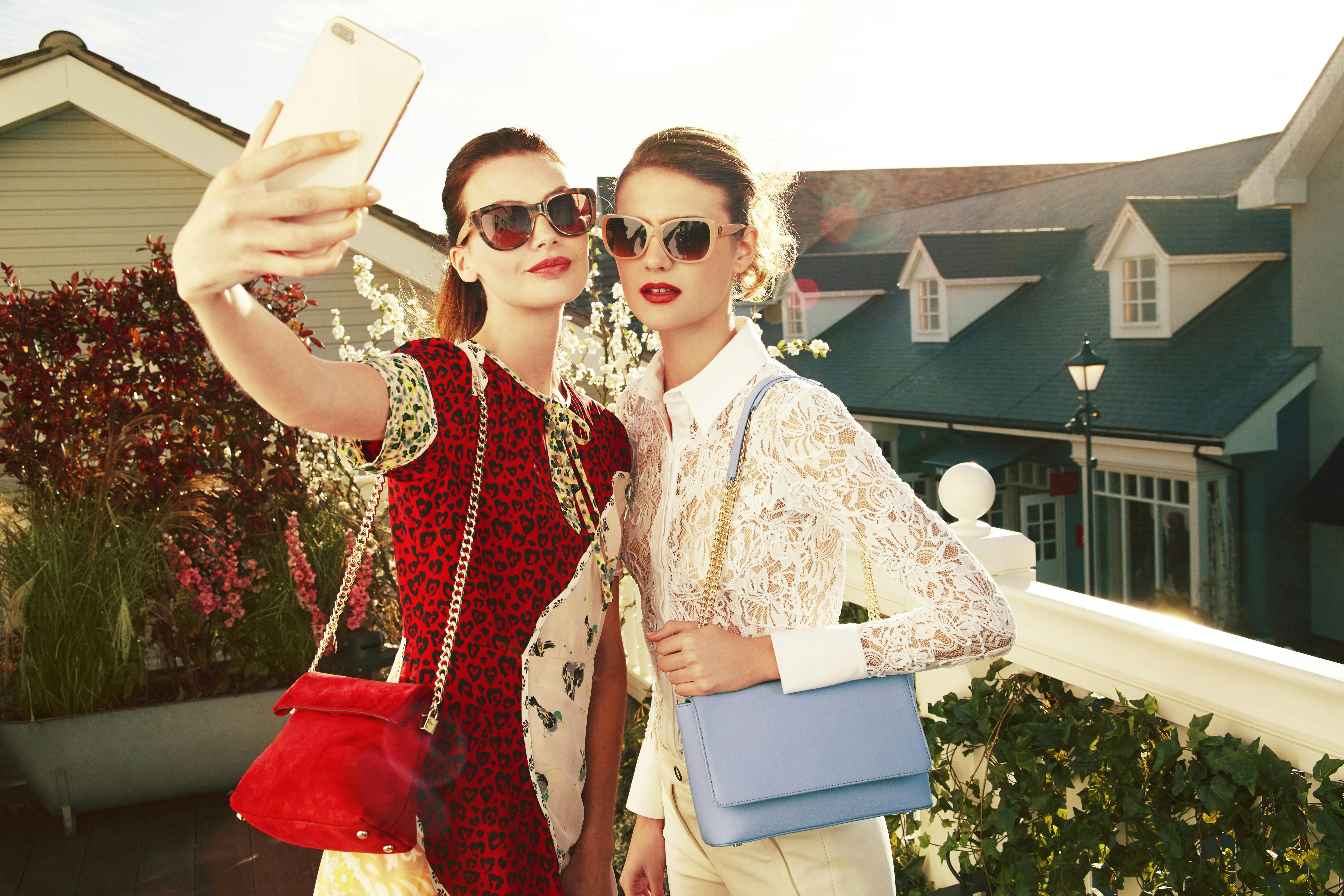 Two women wearing sunglasses and fashionable outfits take a selfie on a sunny terrace with houses in the background.