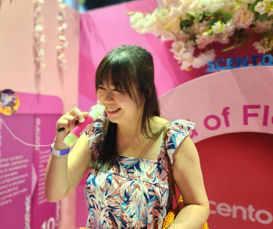 Woman smiles while holding a drink in a vibrant pink environment with floral decor. She wears a colorful, patterned dress.