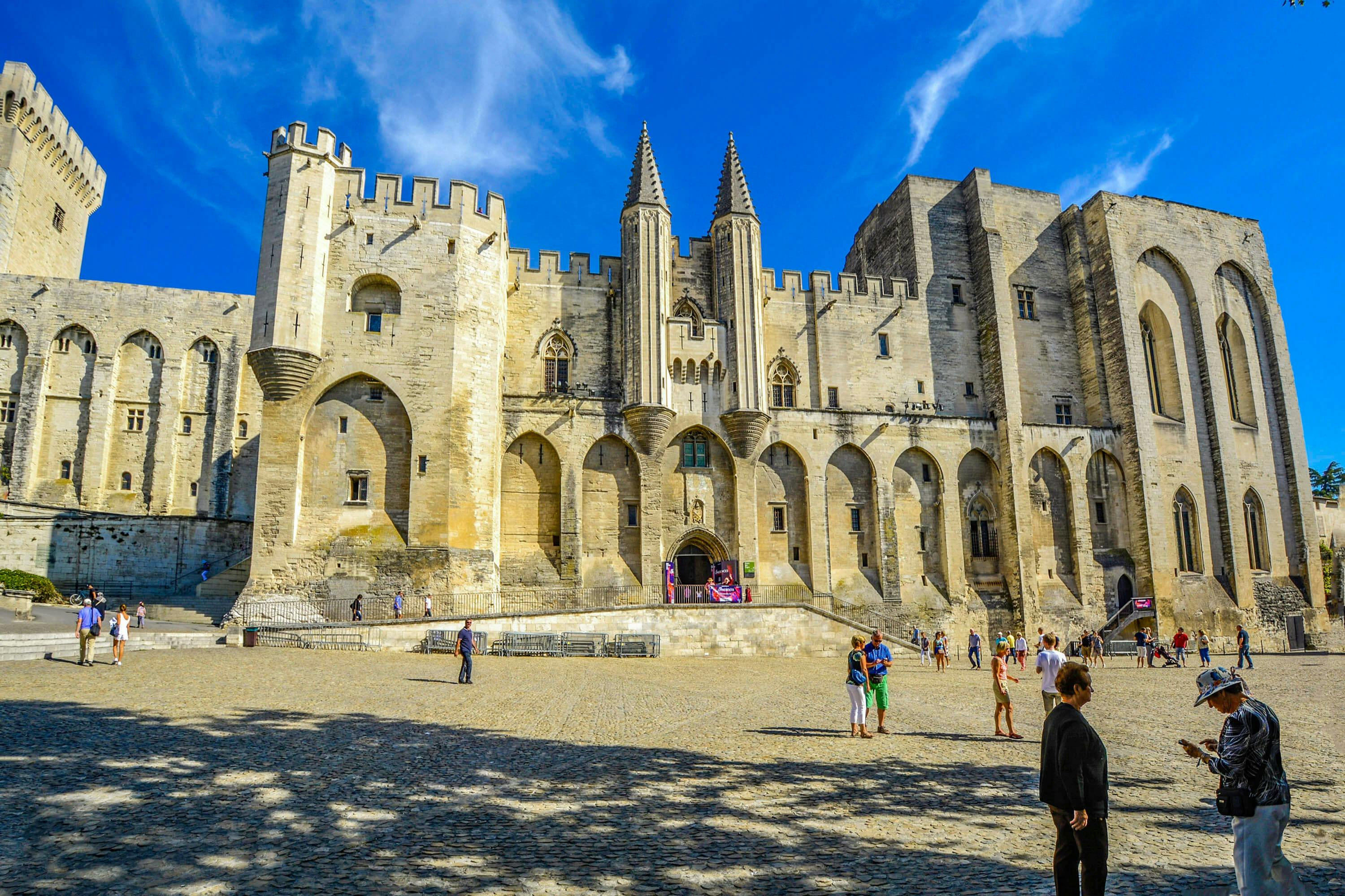 People walking in front of a large, historic stone building with towers and arched windows under a bright blue sky.