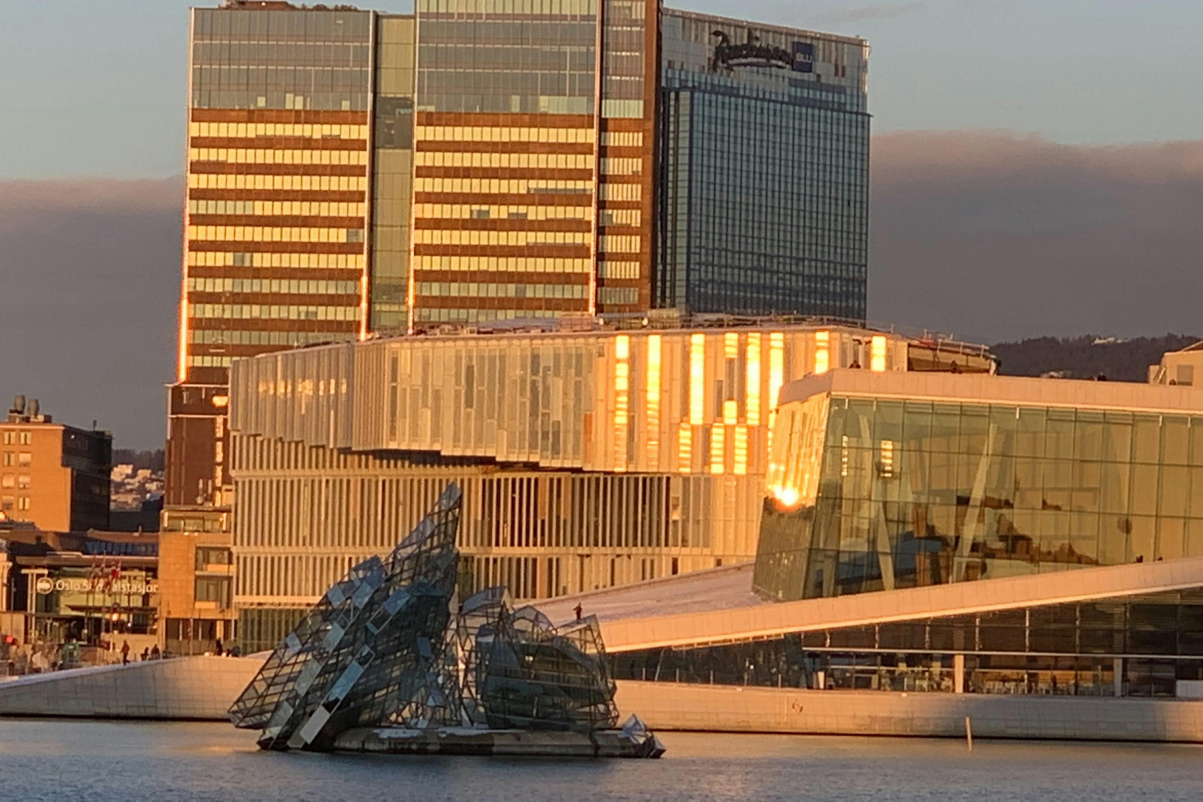 A modern building with reflective glass panels near a waterfront, featuring a glass sculpture in the water under warm light.