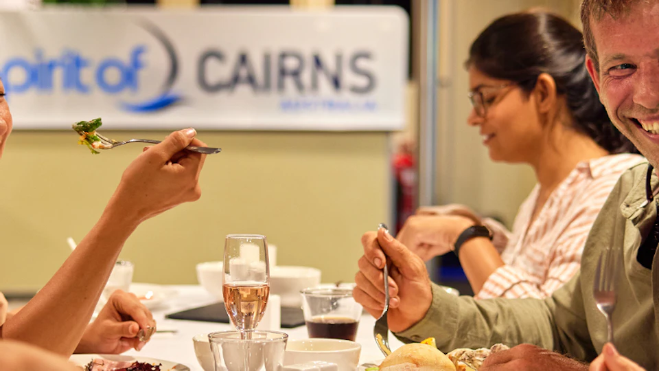 A group of people enjoying a meal together at a restaurant, with a man smiling at a child. A sign reading "CAIRNS" is in the background.