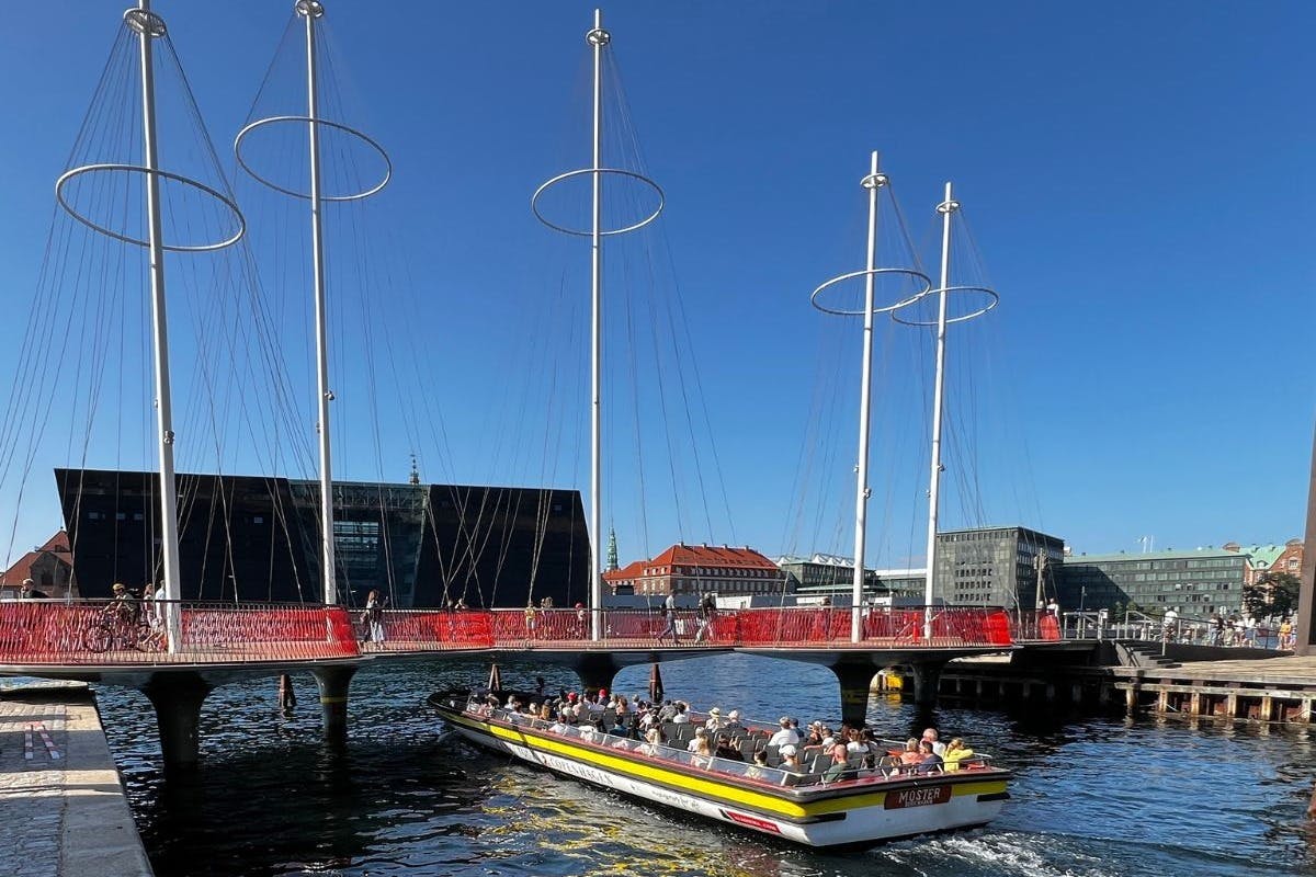 A boat with passengers passes under a modern bridge with circular designs and red railings in a cityscape.