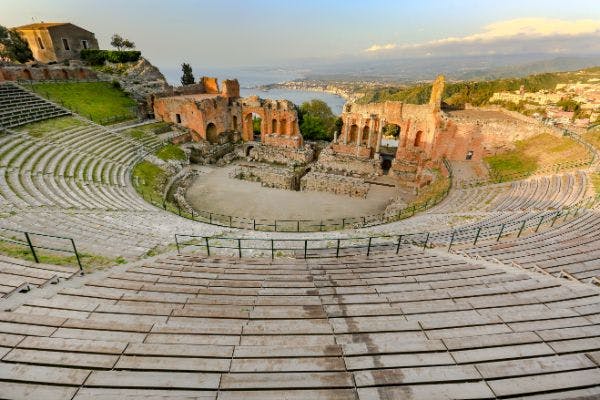 An ancient amphitheater with stone seating, overlooking ruins and a distant coastal landscape under a partly cloudy sky.