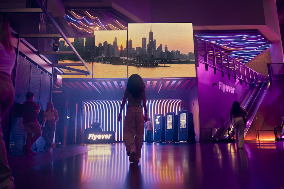 A person walking towards a display featuring a cityscape at sunset, with illuminated signage and decor around.