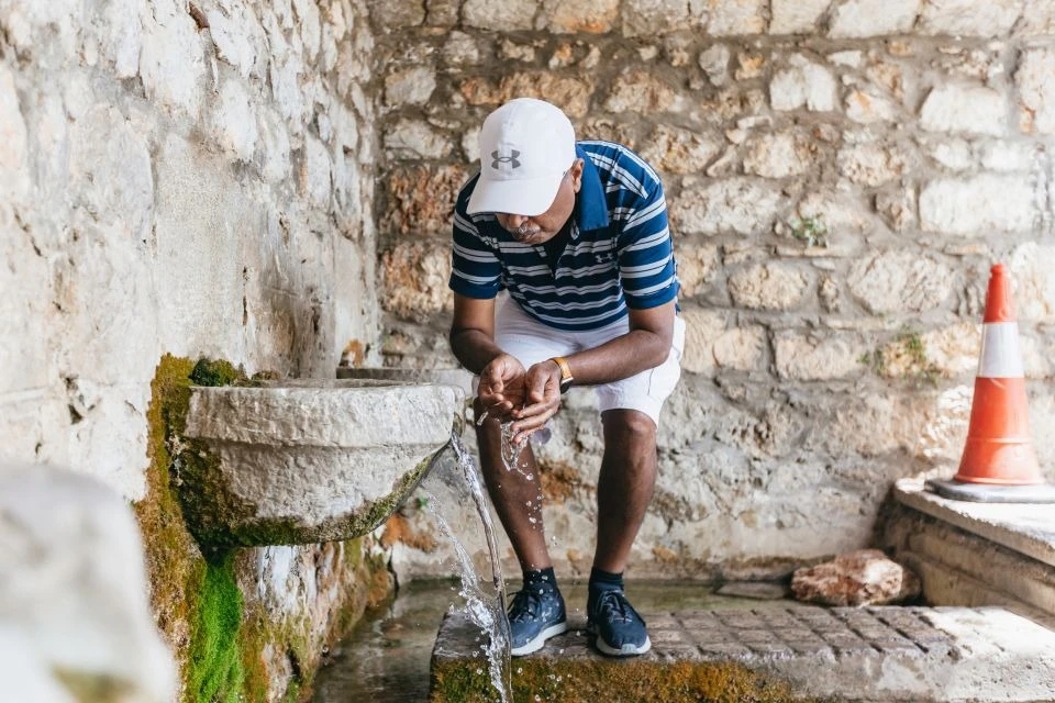 Person in a striped shirt and white cap drinking water from an old stone fountain against a textured stone wall.