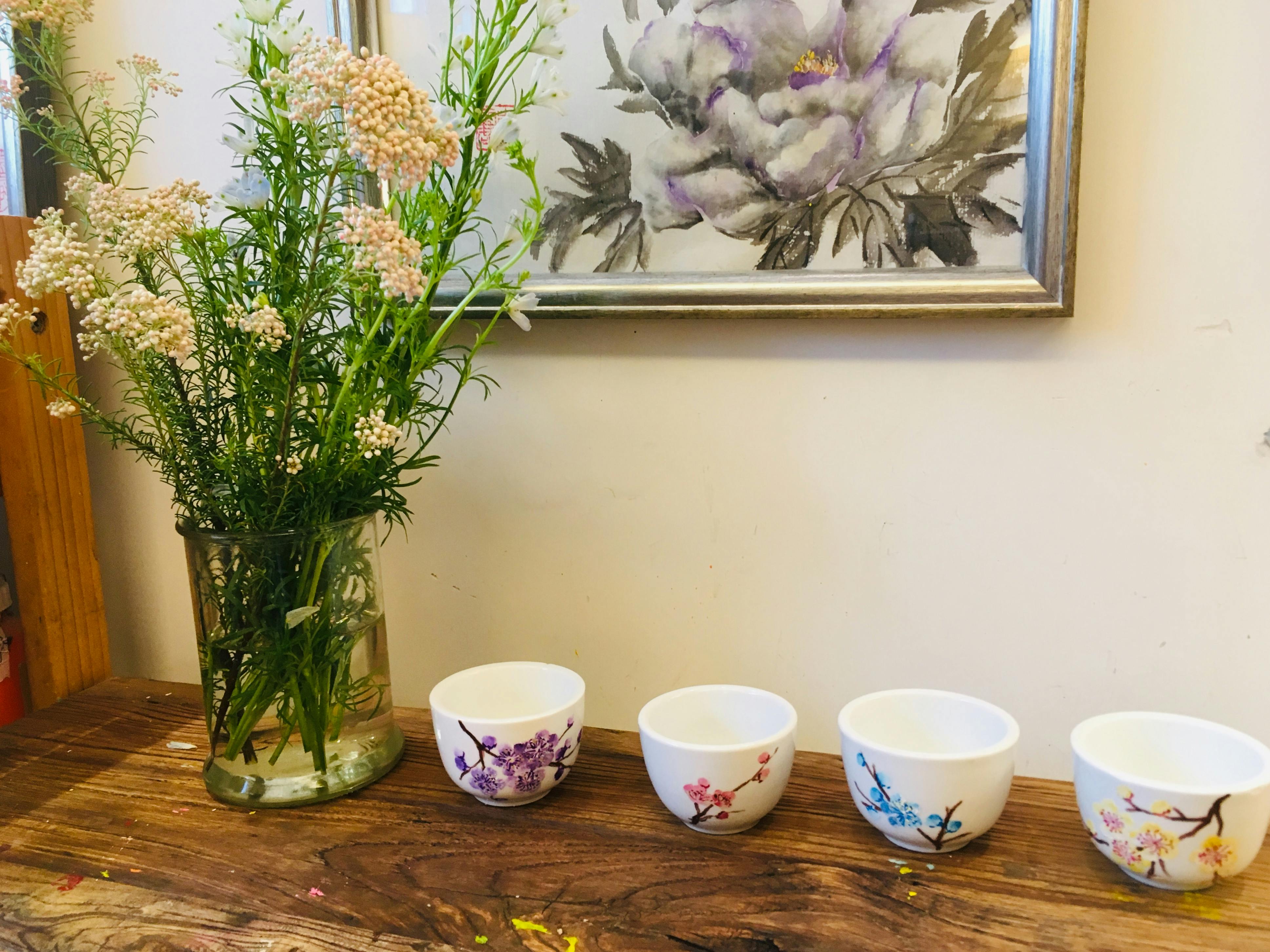 木桌上有四个花卉图案的杯子，旁边是一个花瓶，背景是一幅花卉画。