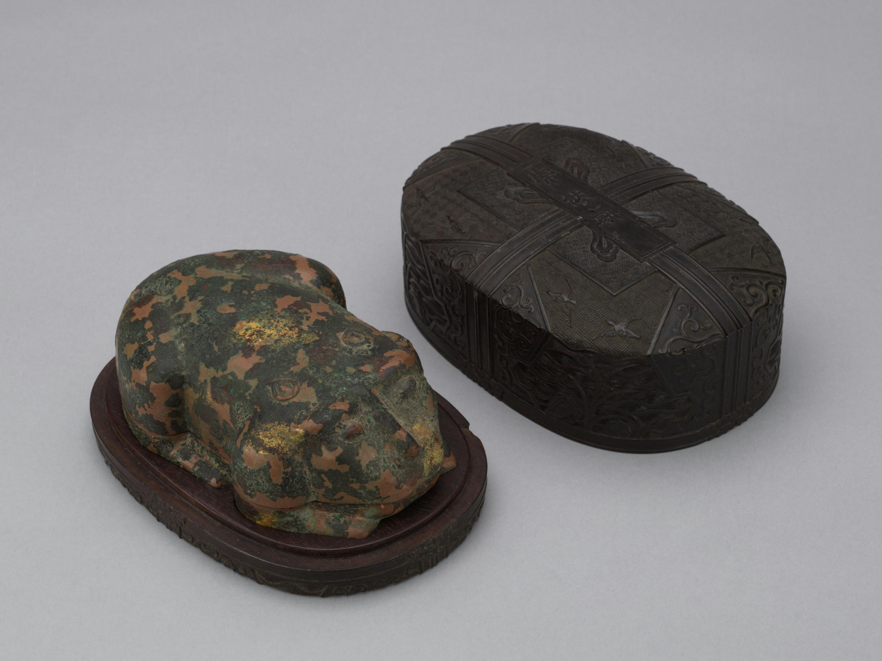 A camouflaged frog-shaped object next to an intricately designed dark oval box on a neutral background.