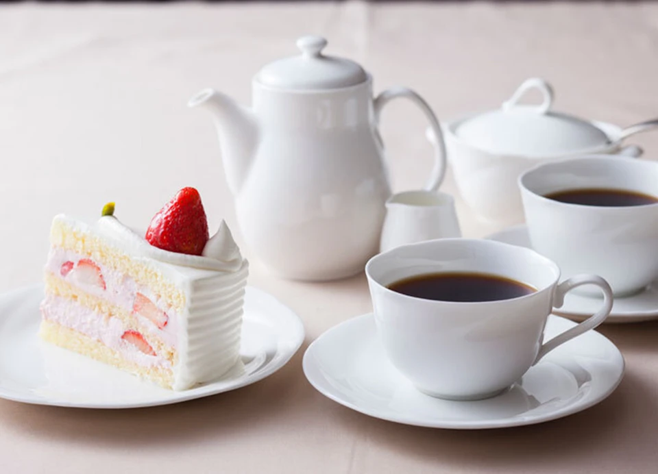 A slice of strawberry shortcake on a plate with a teapot, creamer, and cups of coffee on a table.