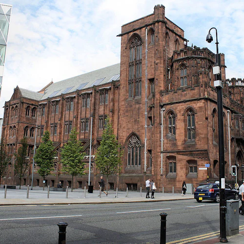 En historisk röd tegelbyggnad på en stadsgata, med fotgängare och bilar. Modern glasbyggnad synlig till vänster.