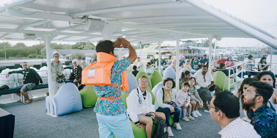 A group of people seated on cushions under a canopy on a boat, listening to a man in a life vest addressing them.
