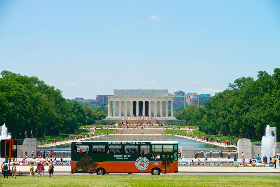 A double-decker tour bus in front of the Lincoln Memorial surrounded by people and greenery on a clear day.