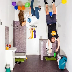 Två personer visas upp och ner i ett badrum med färgglada ballonger på golvet, vilket skapar en optisk illusion.