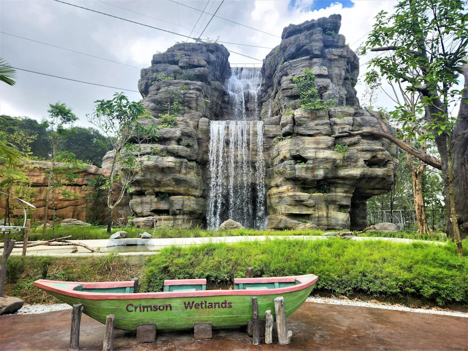 A green and red boat labeled "Crimson Wetlands" in front of a large rock formation with a waterfall and greenery.