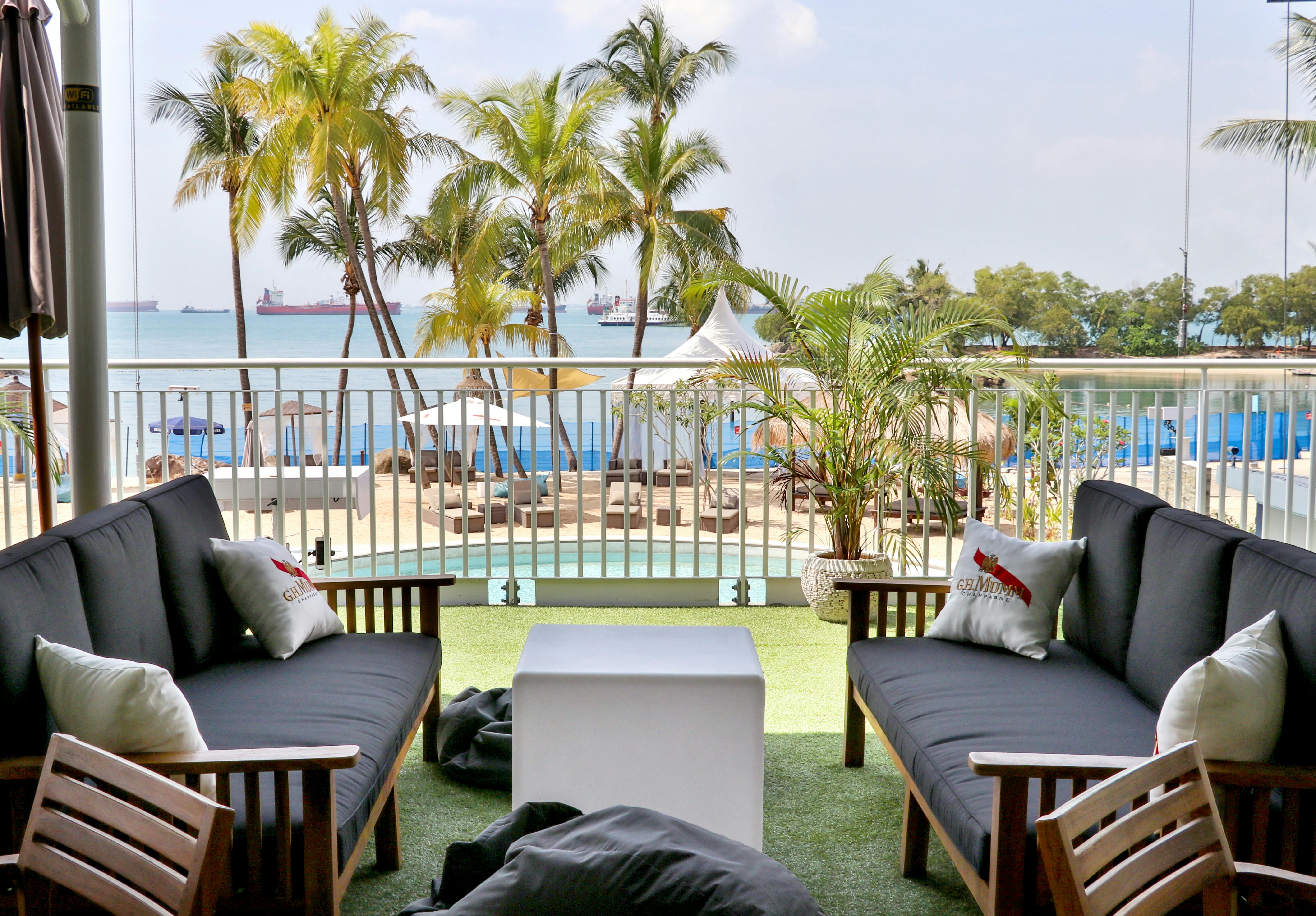Two lounge chairs face a small table on a balcony overlooking a pool, palm trees, and the ocean with ships in the distance.