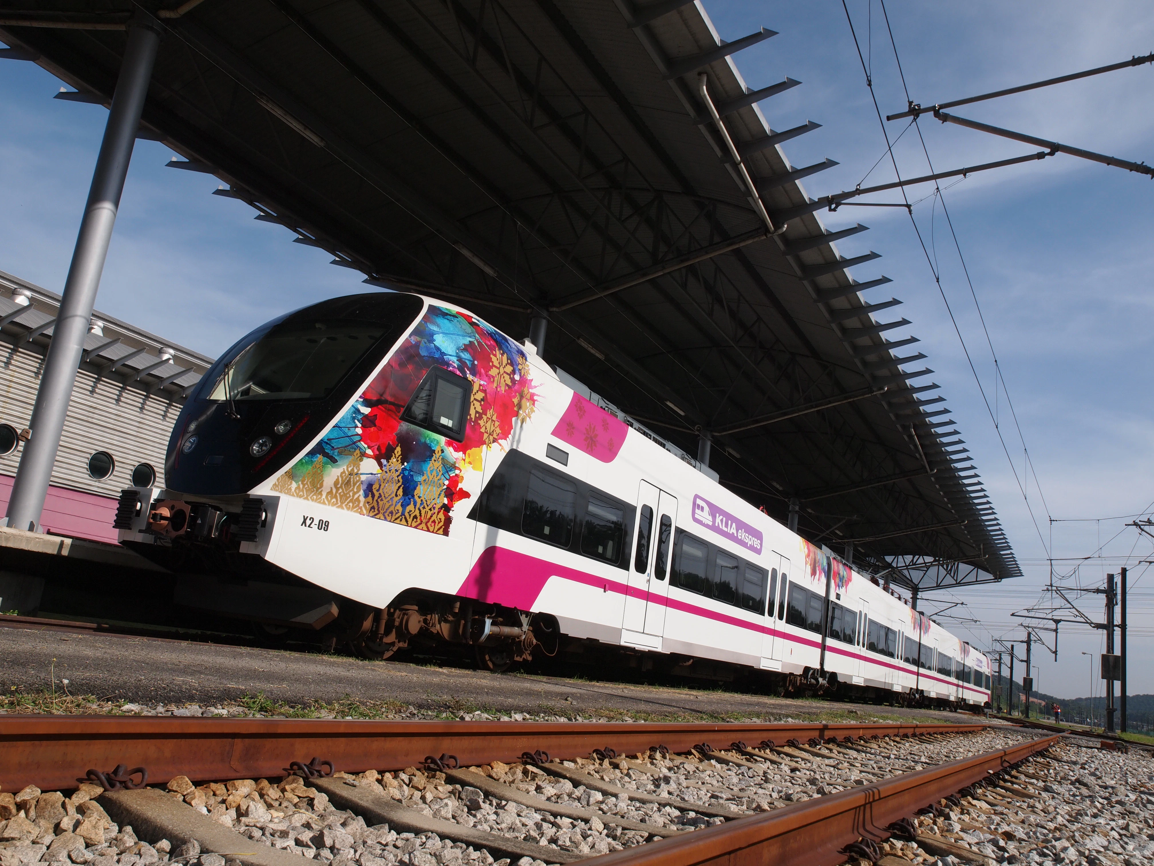 A white train with colorful floral designs and a "KLIA Ekspres" sign is stationary under a metal canopy at a railway station.