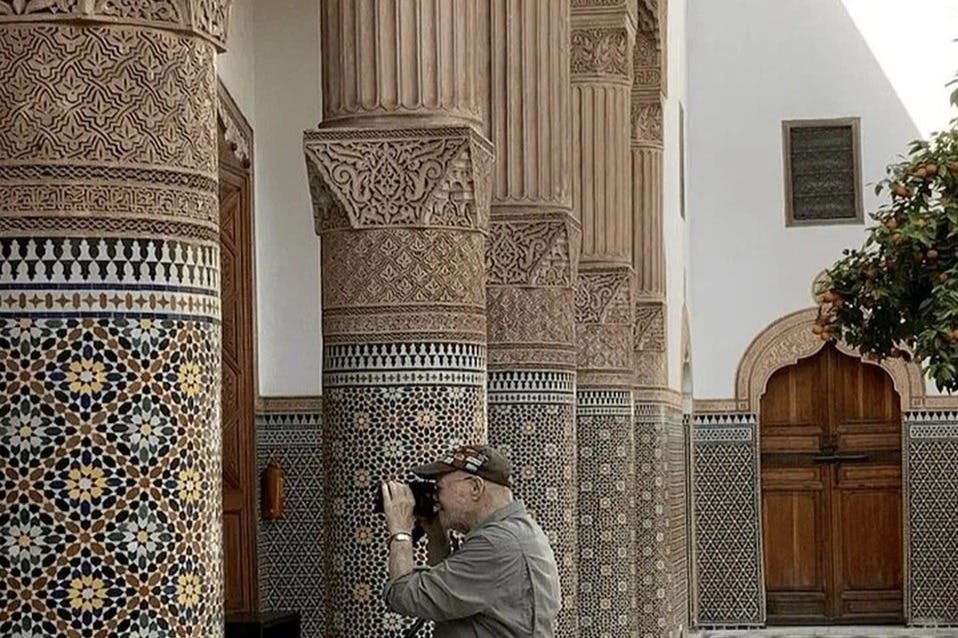 A person with a camera takes pictures of intricately decorated columns in a tiled and ornate building interior.