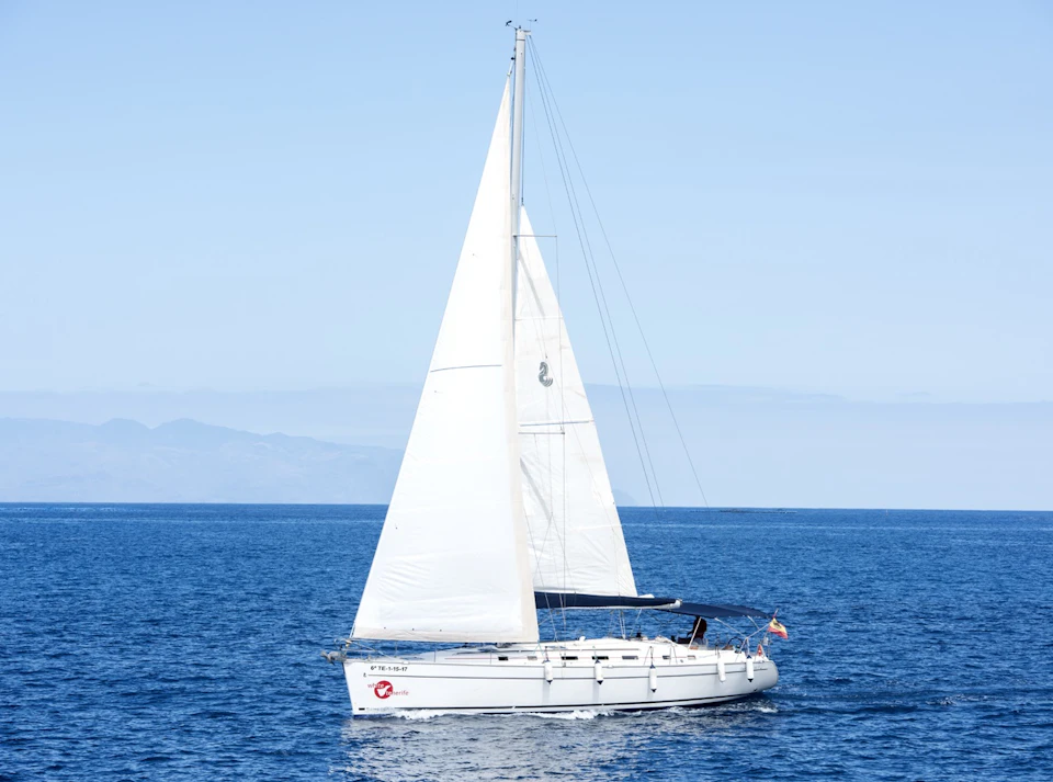 A white sailboat with large sails glides on a calm, blue ocean under a clear sky with distant mountains on the horizon.