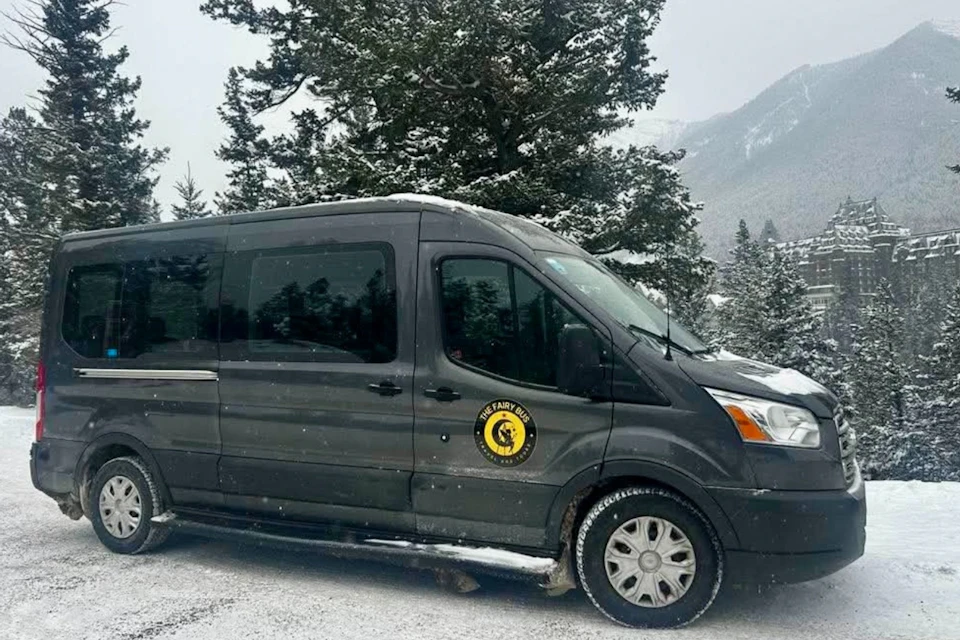 A black van with a logo parked on a snowy road, surrounded by snow-covered trees and mountains in the background.