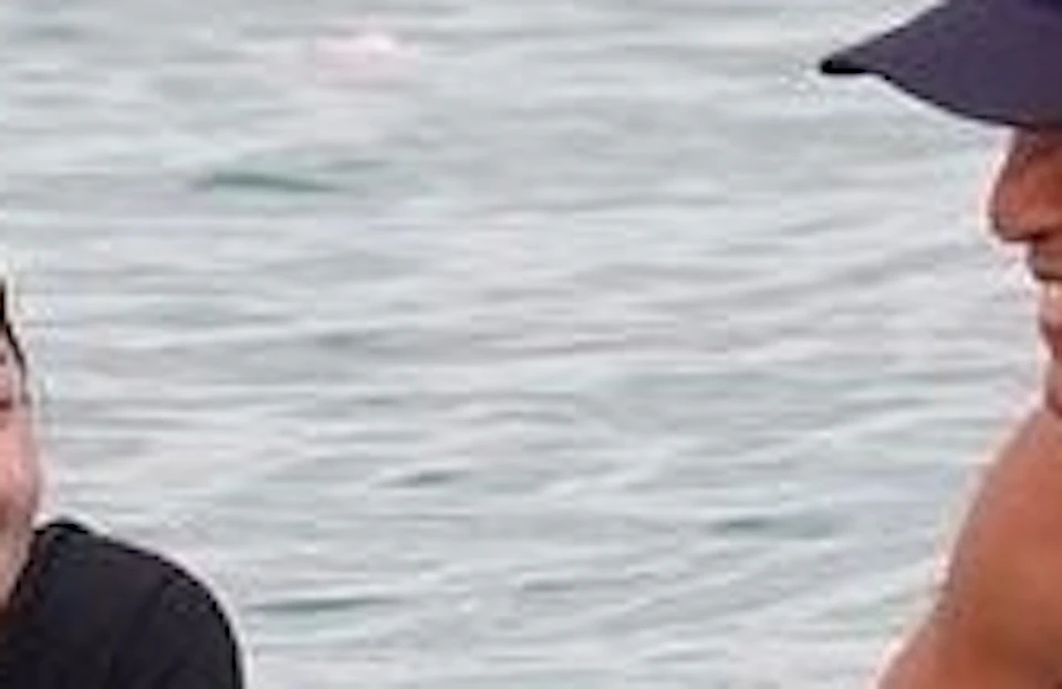 Two people on surfboards in the water, one smiling and lying on a board, the other standing and holding another board.