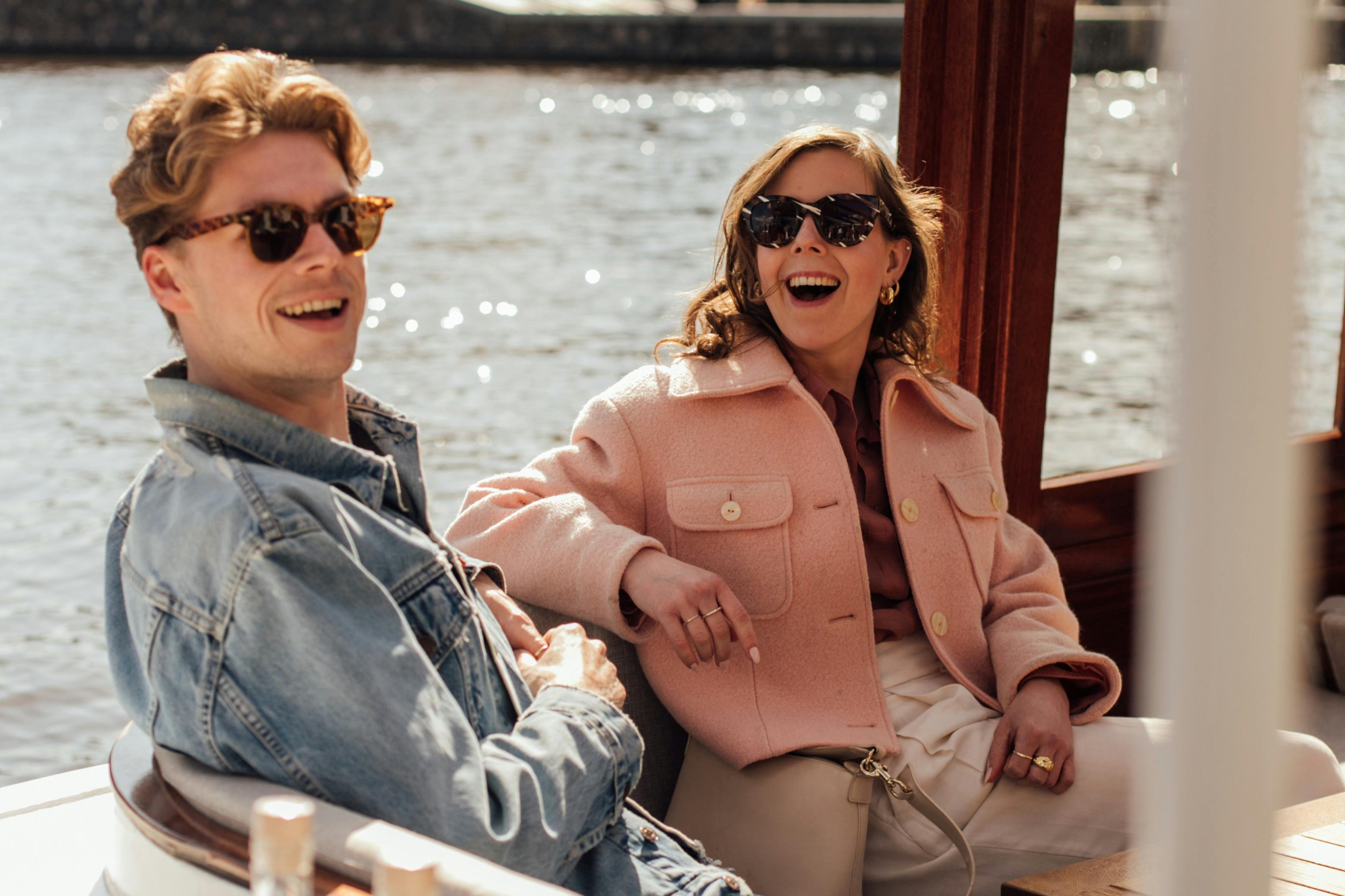 Deux personnes assises sur un bateau, souriantes et portant des lunettes de soleil, avec de l'eau et un rivage lointain en arrière-plan.