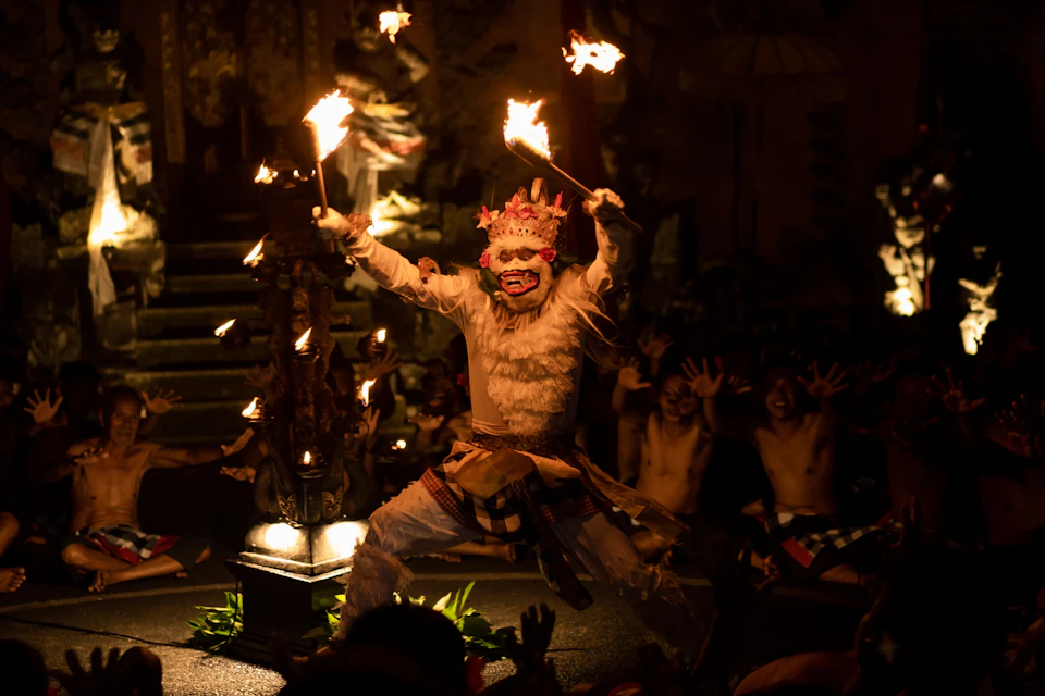 A performer in traditional costume and mask holding flaming torches during a nighttime cultural performance with an audience.