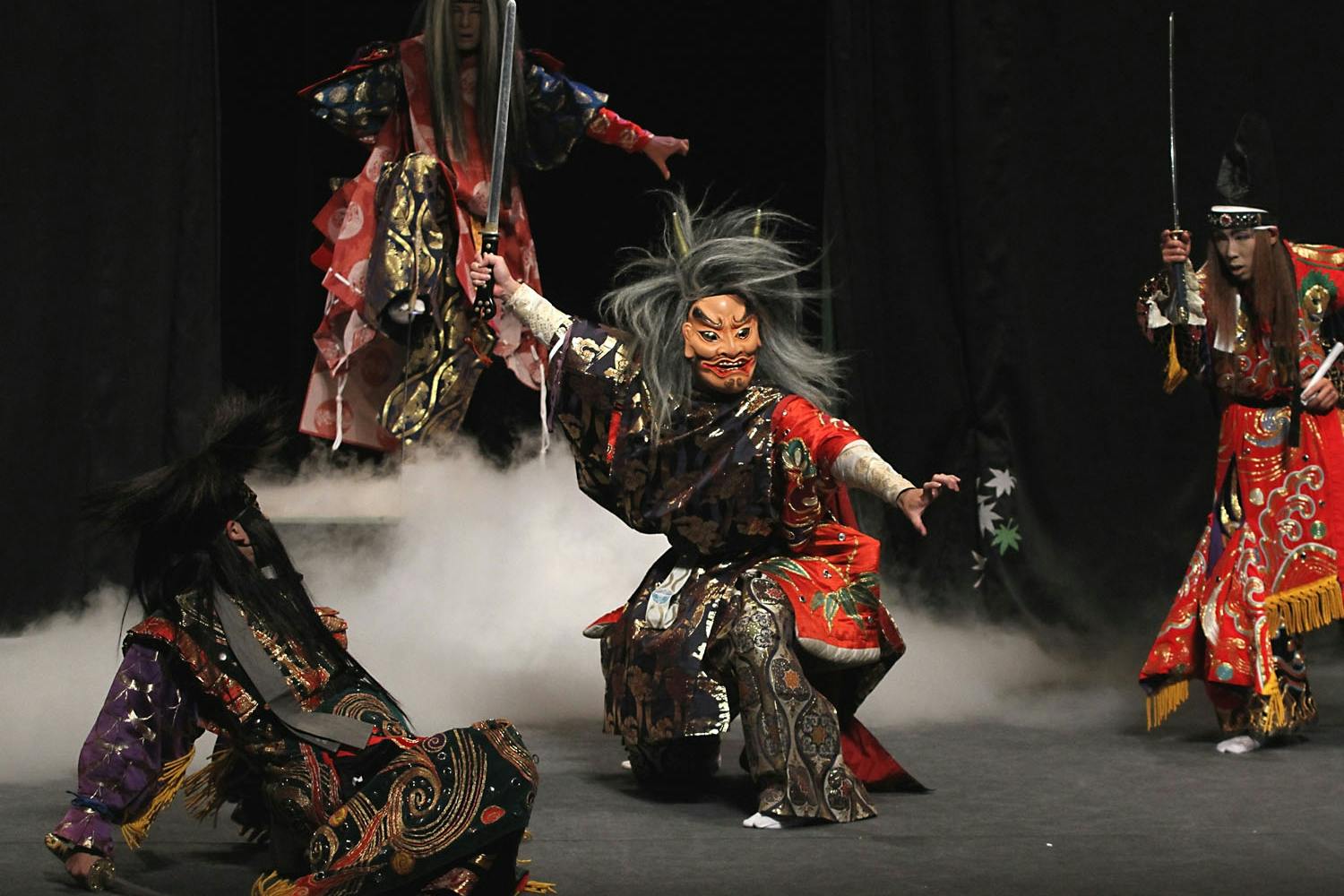Performers dressed in traditional Japanese costumes and masks enact a dramatic scene on stage amidst smoke effects.