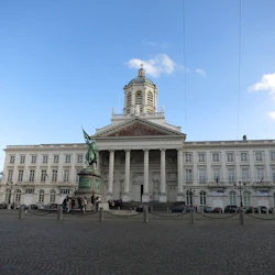 Storslagen byggnad med ett centralt klocktorn, pelare vid ingången och en staty av en monterad figur framför på en kullerstensplaza.