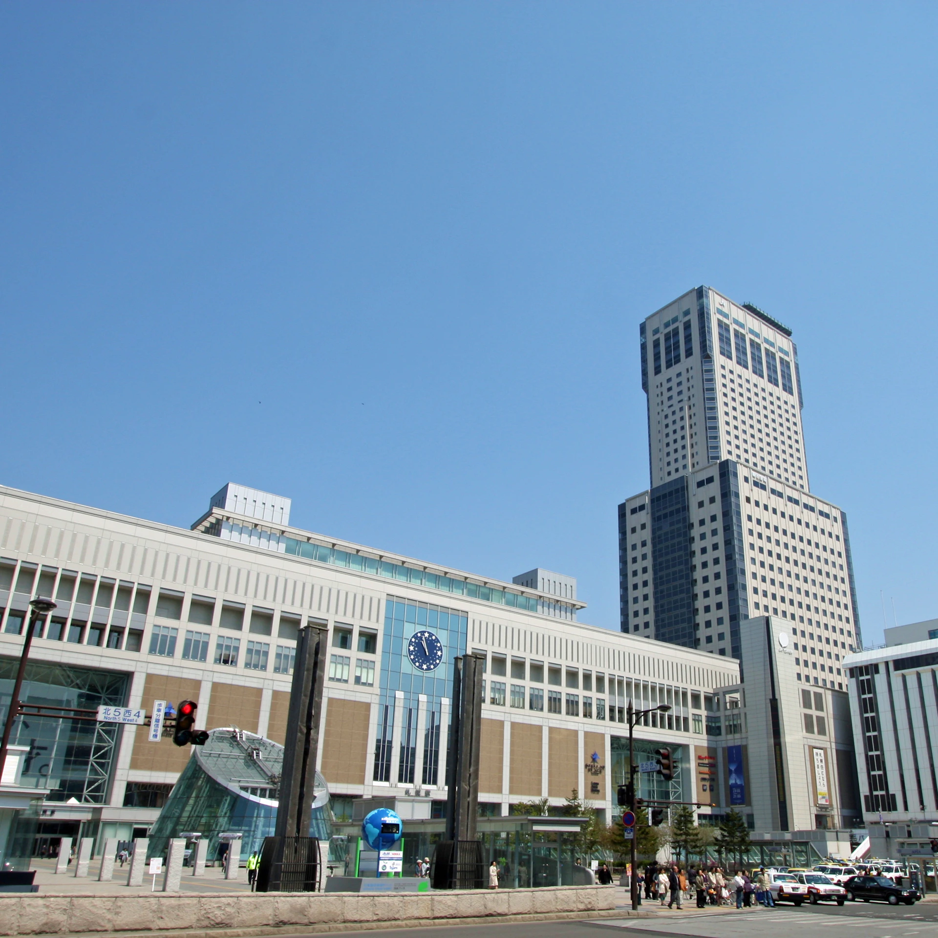 Modern buildings with large windows and a clock on the facade under a clear blue sky. People and cars are visible at the street level.