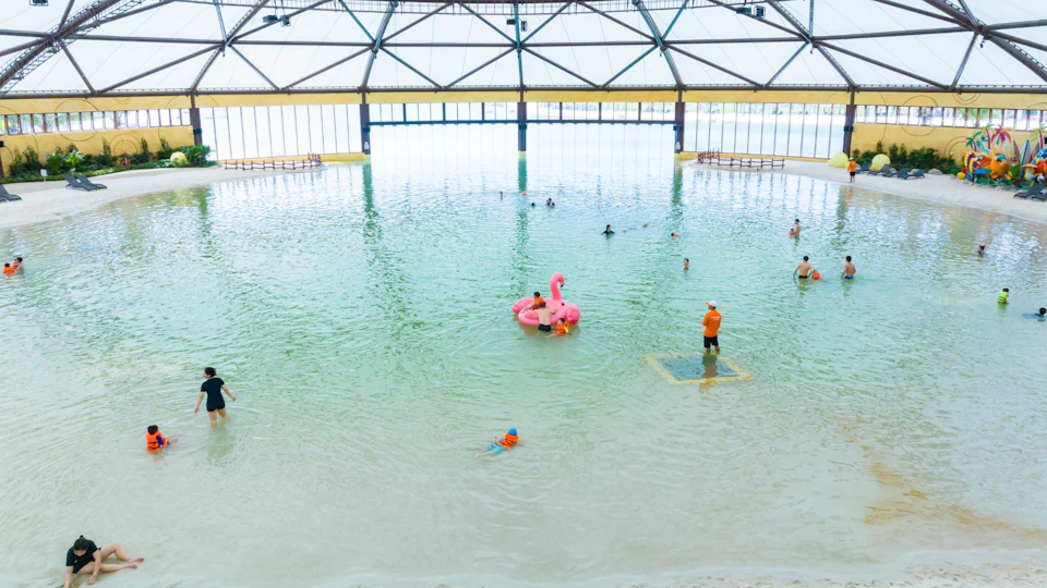 People enjoy a large indoor pool with a pink flamingo float, surrounded by a high, arched, and transparent roof.