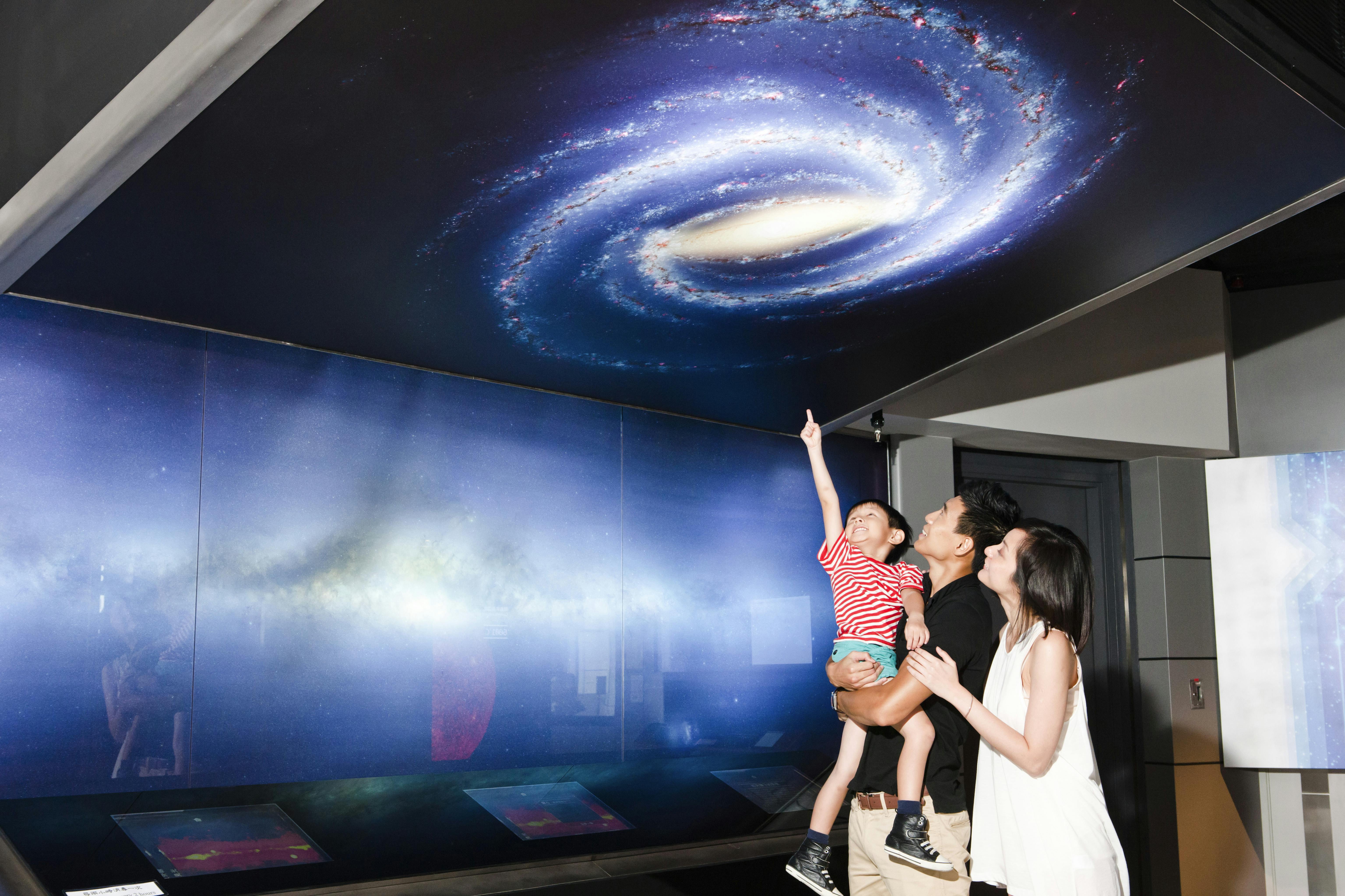 Семья смотрит на большой детальный галактический дисплей на выставке; ребенок взволнованно указывает на закрученные огни.