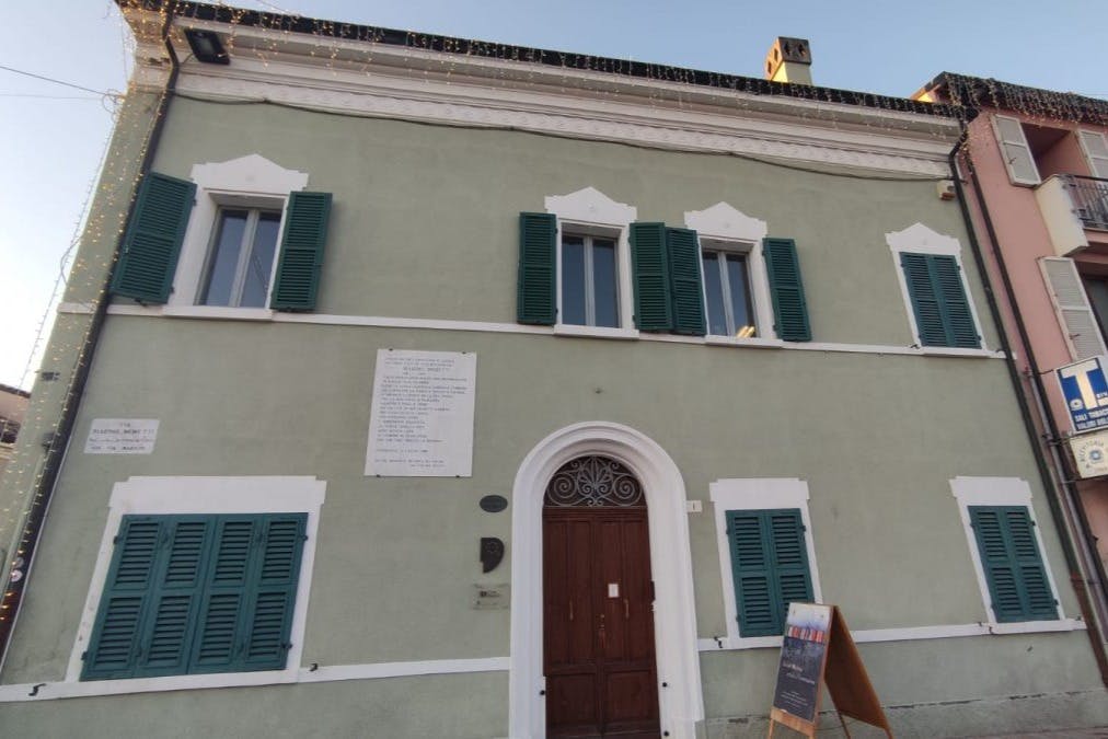 Un edificio verde chiaro con rifiniture bianche, persiane verdi e una doppia porta in legno. Sono visibili una targa e un'insegna con lavagna.