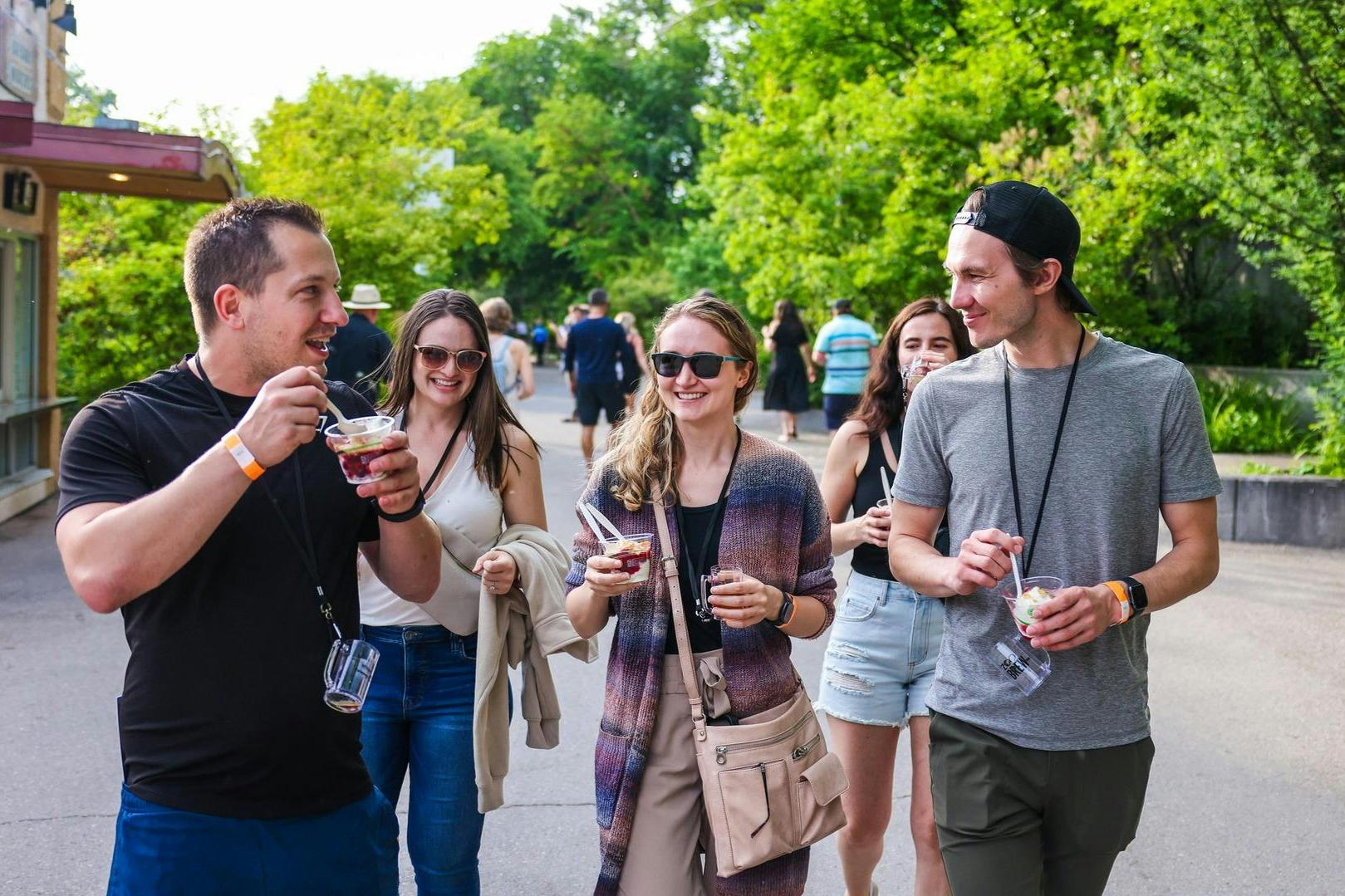 Skupina lidí, někteří se slunečními brýlemi, se prochází venku a za slunečného dne si vychutnává jídlo z šálků. V pozadí jsou stromy.
