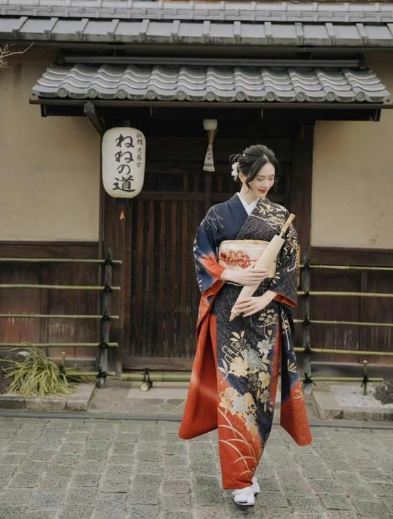 A woman in a traditional Japanese kimono stands in front of a wooden gate with a sign and tiled roof.