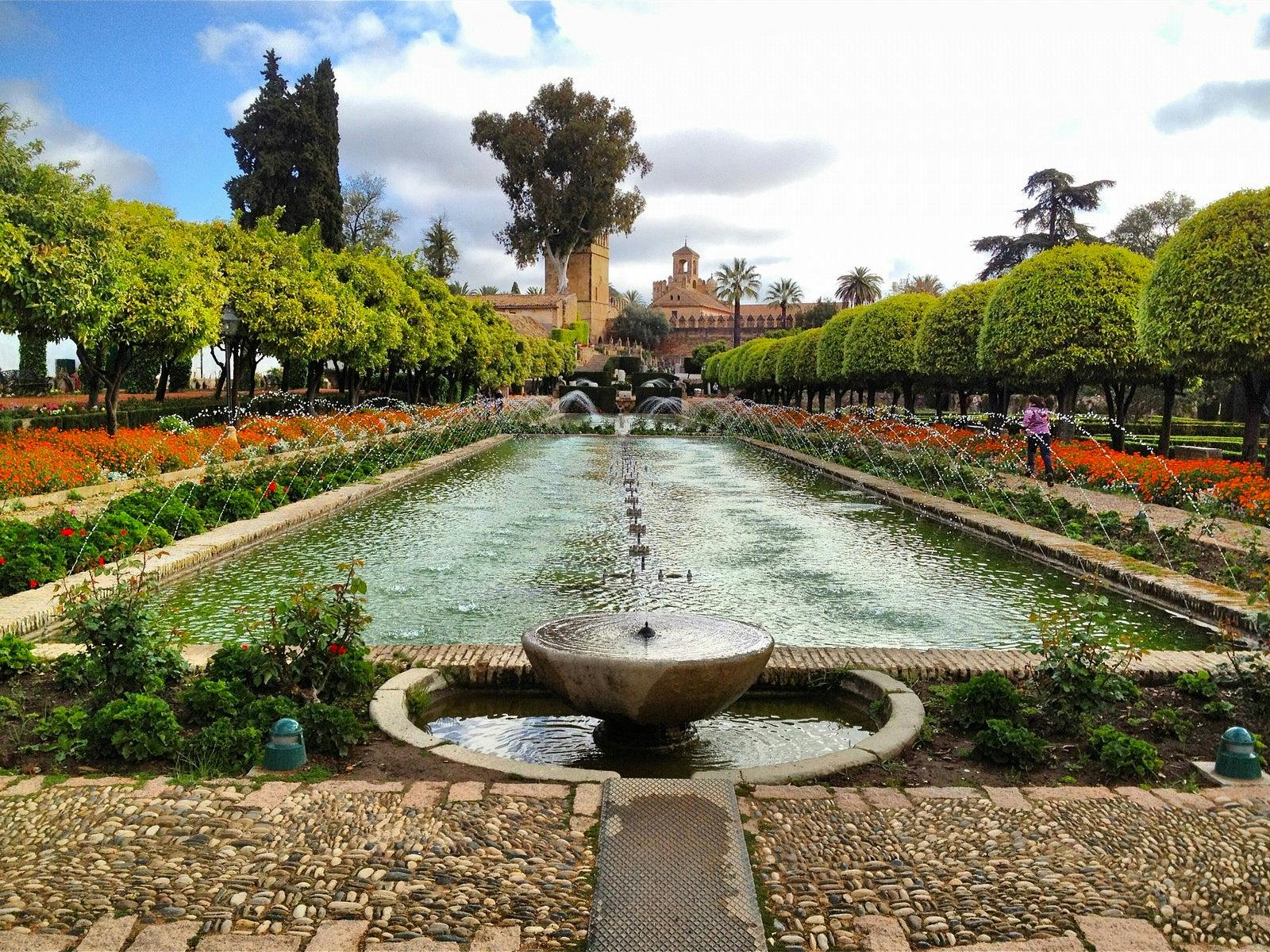 A long fountain surrounded by trees and flowers leads to an ornate building under a partly cloudy sky.