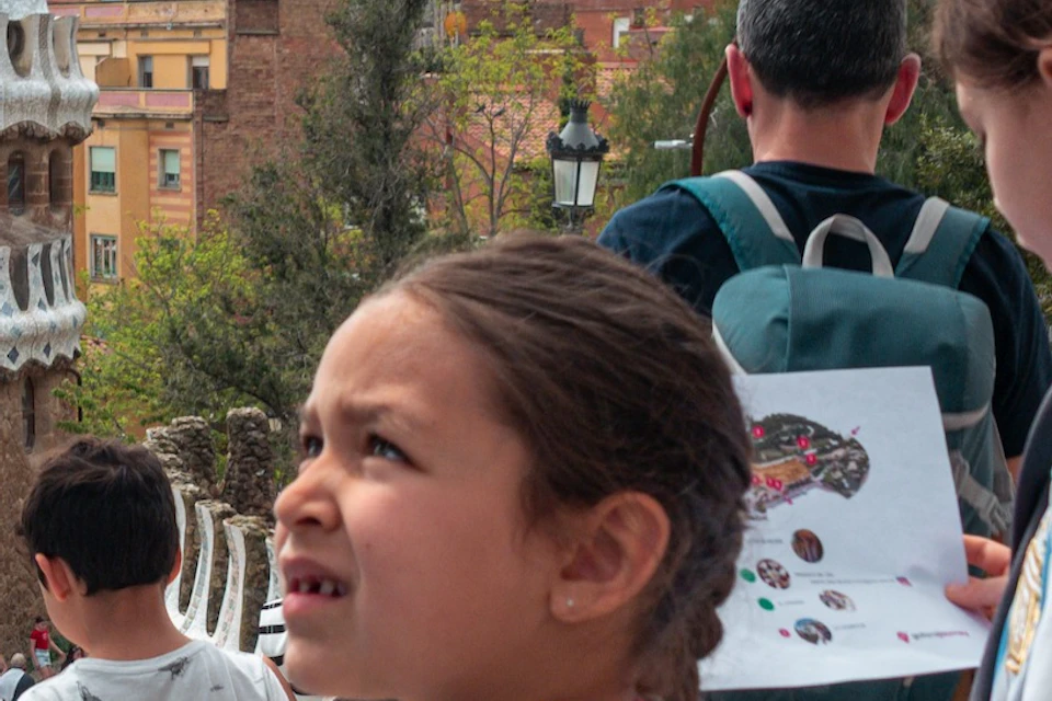Children holding maps explore an outdoor area near architectural structures with unique designs and a city backdrop.