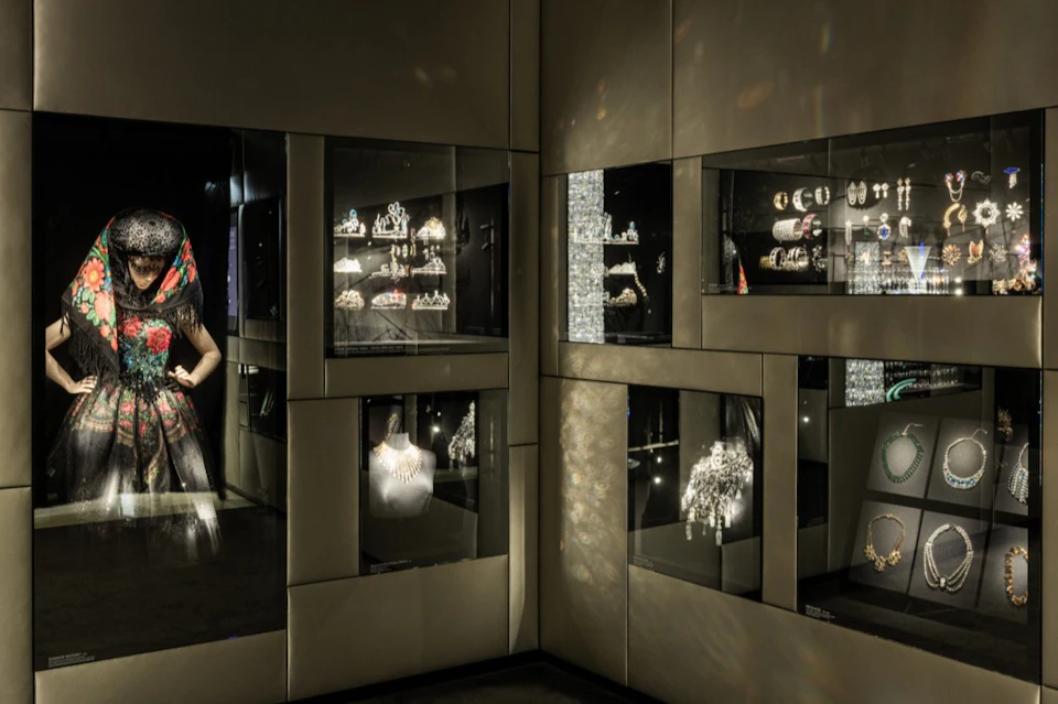 Dimly lit display of jewelry and accessories in a museum-like setting. A mannequin wears a colorful scarf.