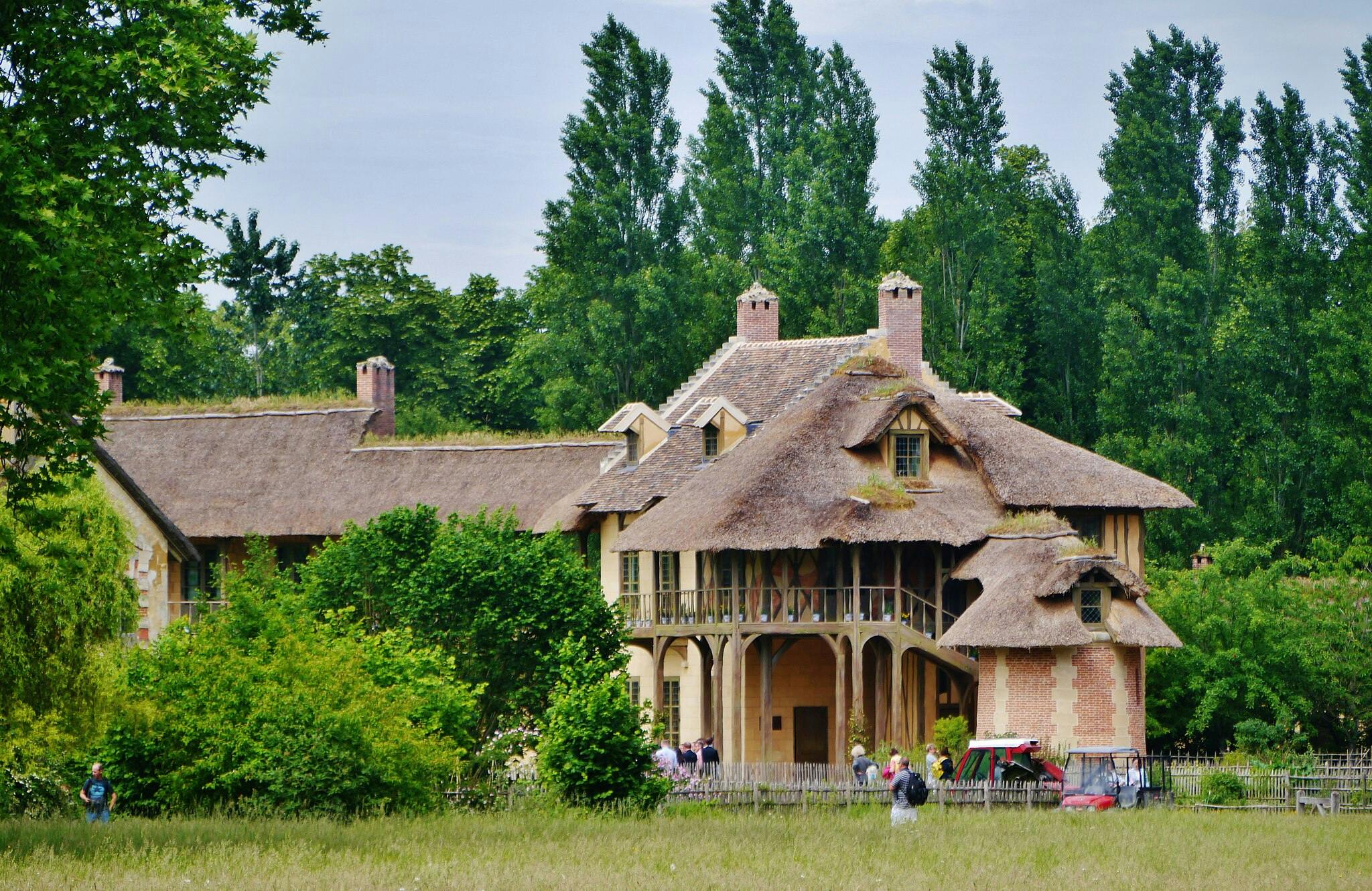 Chalupa s doškovou střechou se dvěma komíny, obklopená zelenými stromy a keři. V blízkosti budovy stojí skupina lidí.