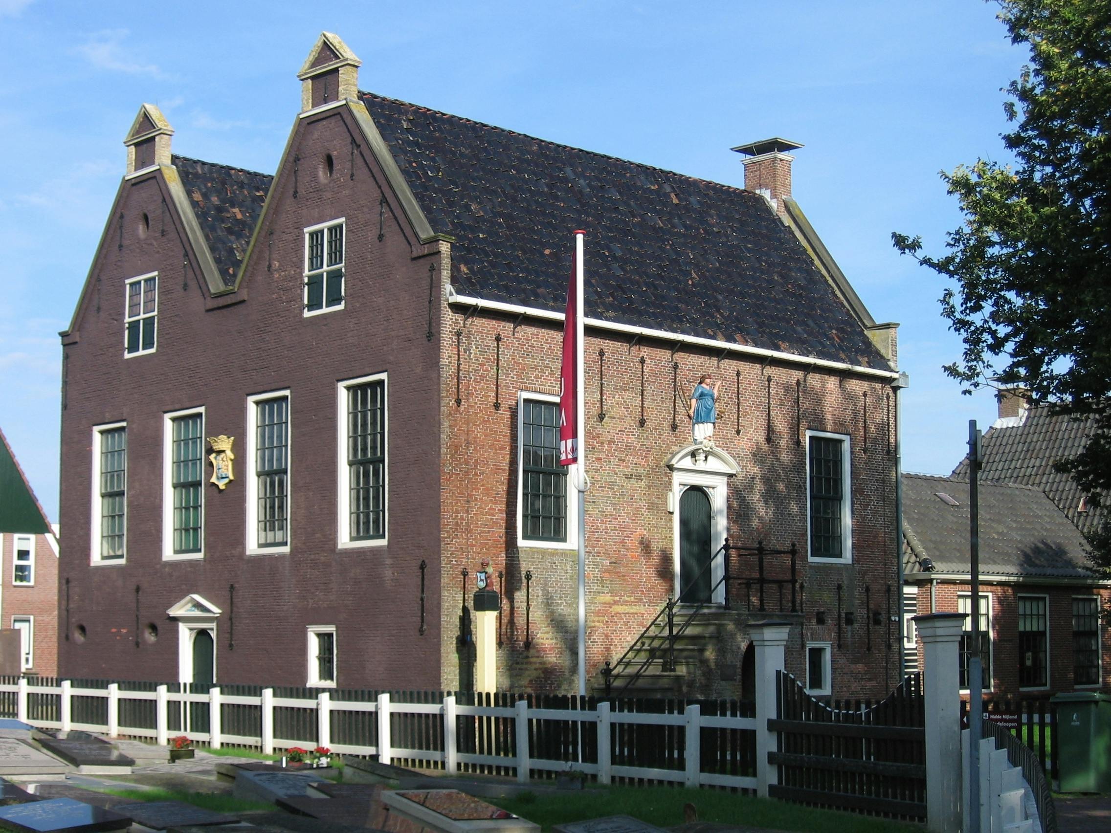 Een historisch bakstenen gebouw met grote ramen, een trap die naar een verhoogde ingang leidt en een omheining van wit piket.