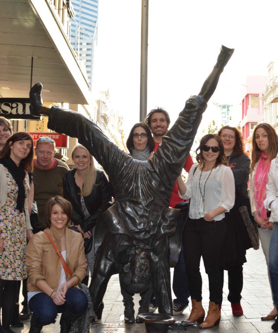 도심의 거리를 배경으로 한 남성이 손을 잡고 서 있는 청동 동상 주위에서 포즈를 취하고 있습니다.