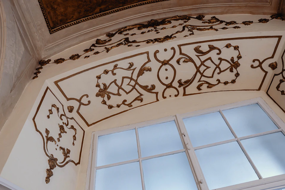 Ornate ceiling with gold decorative patterns above a window with blue sky visible.