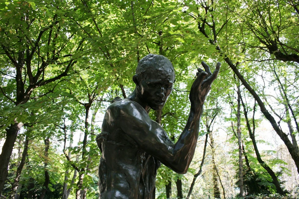 A bronze statue of a man with an outstretched arm set against a backdrop of lush, green trees.