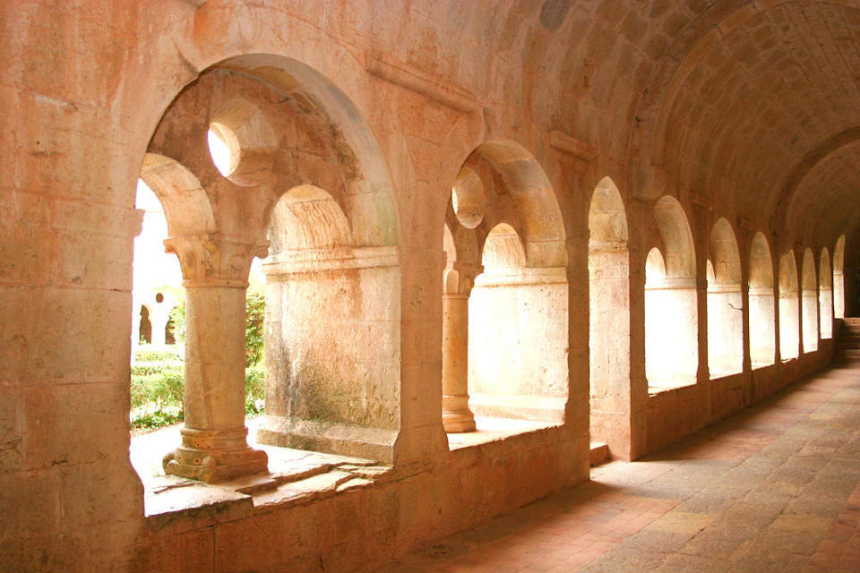 Stone walkway with arched openings, columns, and a sunlit courtyard with greenery in the background.