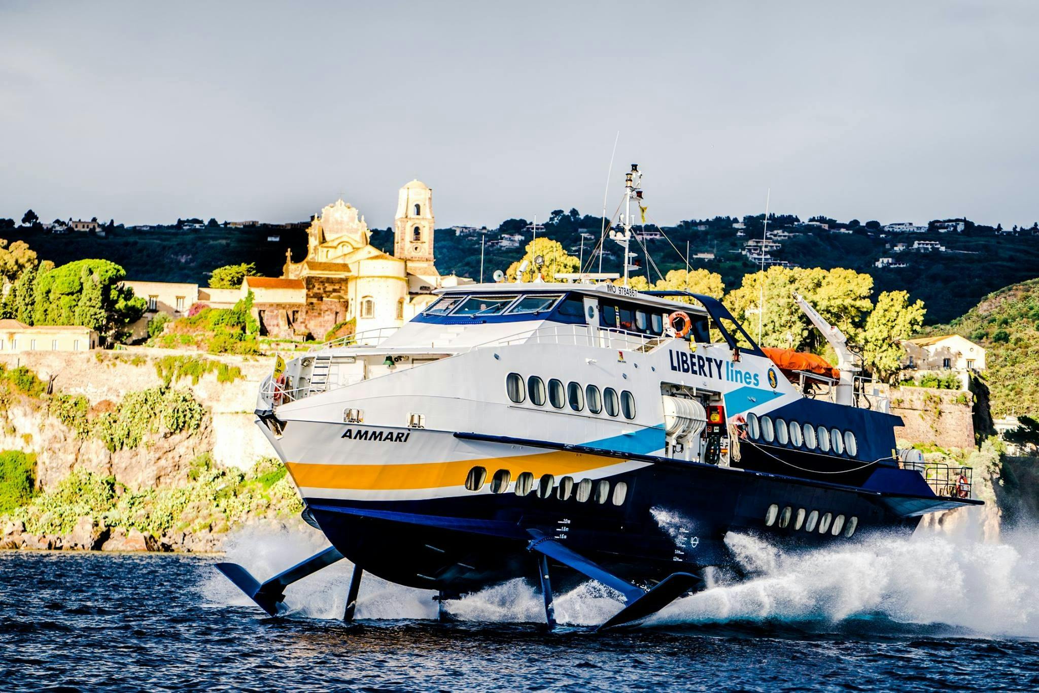 A hydrofoil boat named "Ammari" by Liberty Lines moves swiftly on water, with a hillside village and buildings in the background.
